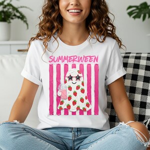 May include: White t-shirt with the word "SUMMERWEEN" in pink above a ghost illustration. The ghost wears sunglasses and is covered in strawberries, holding a pink drink. Pink vertical stripes are behind the ghost.