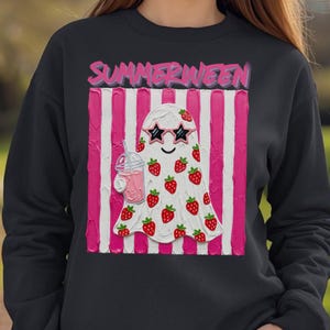 May include: Black sweatshirt featuring a white ghost with strawberry accents, star-shaped sunglasses, and a pink drink. The ghost is set against a pink and white striped background, with the word "SUMMERWEEN" in pink above.