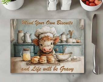 Könnte beinhalten: Rechteckiges Schneidebrett mit einer Aquarellillustration eines Kochs in Kuhform. Die Kuh trägt eine Kochmütze und eine Schürze mit dem Text "Mind Your Own Biscuits and Life will be Gravy". Der Hintergrund zeigt eine Küchenszene.