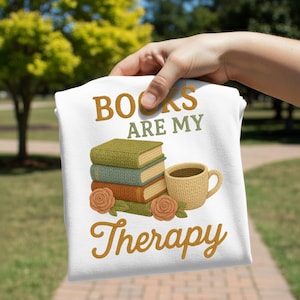 Puede incluir: Camiseta blanca con el texto "BOOKS ARE MY Therapy" en escritura marrón. El diseño incluye una pila de libros, una taza de café y rosas en tonos verdes, marrones y beige. La camiseta está sujeta por una mano.