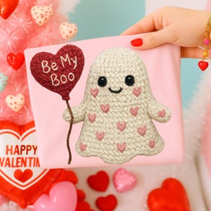 May include: A pink Valentine's Day card with a crocheted ghost holding a heart-shaped balloon that reads "Be My Boo." The ghost has pink heart details and a smiling face. The background is a soft pink.