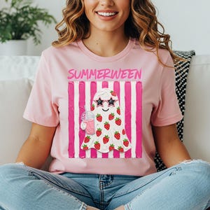 May include: Pink t-shirt with the word "SUMMERWEEN" above a ghost illustration. The ghost wears star-shaped sunglasses and is covered in strawberries. It holds a pink drink with a straw. The background has pink and white vertical stripes.