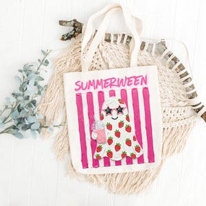 May include: A white canvas tote bag with the word "SUMMERWEEN" in pink letters. The bag features a ghost illustration wearing star-shaped sunglasses and covered in strawberries, holding a pink drink. Pink vertical stripes are behind the ghost.