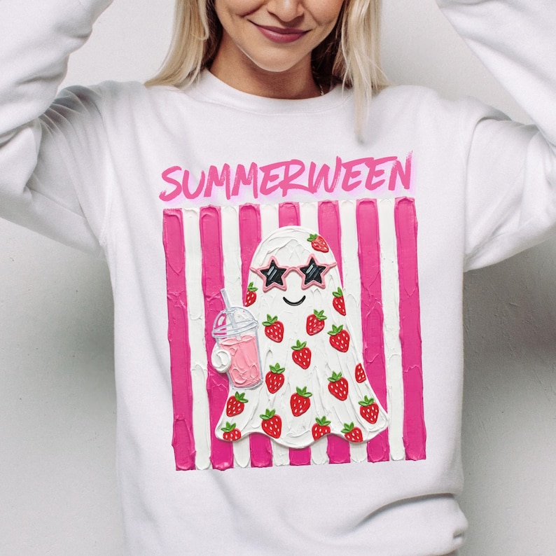 May include: White sweatshirt with the word "SUMMERWEEN" in pink. The design features a ghost wearing star-shaped sunglasses and covered in strawberries, holding a pink drink. Pink and white striped background.