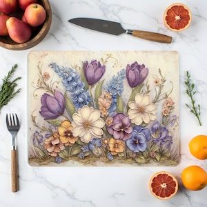 Könnte beinhalten: Rechteckiges Schneidebrett mit floralem Design mit lila Tulpen, blauen Hyazinthen und weißen und lila Stiefmütterchen. Eine Holzschale mit Äpfeln, ein Messer und Orangen sind um das Brett angeordnet.