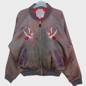 May include: A brown bomber jacket with embroidered bird designs. The jacket features a red lining, green and red striped trim on the collar, cuffs, and hem, and purple pocket accents. It has a full zipper closure and is hanging on a hanger.