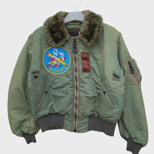 May include: An olive green bomber jacket featuring a faux fur collar, a central zipper closure, and ribbed cuffs and hem. A circular patch on the chest displays a yellow airplane graphic with the text MOSQUITO, alongside a brown rectangular patch.