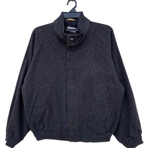 May include: A dark grey wool jacket with a high collar and two front pockets. The jacket has a button closure and is hanging on a black hanger. The jacket's design is classic and suitable for cooler weather.