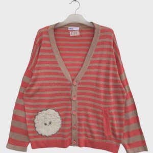 May include: A V-neck cardigan featuring horizontal stripes in coral pink and beige. The garment has a button-down front with matching coral pink buttons and a white, textured, circular patch with a small face design on the lower left side.