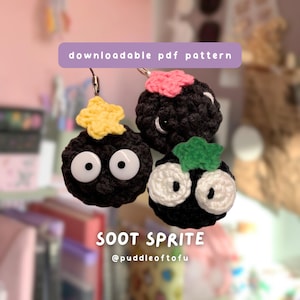 May include: Three crocheted Soot Sprite keychains. Each black sphere has large white eyes and a small decorative element on top, including a yellow star, pink flower, and green leaf. The text "downloadable pdf pattern" is above the sprites.