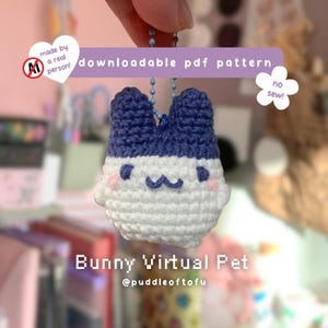 May include: A small, crocheted bunny charm featuring a white body and dark blue ears. The face has embroidered dark blue eyes, a curved mouth, and pink cheeks. It hangs from a blue ball chain. Text includes "downloadable pdf pattern", "no sew!", and "Bunny Virtual Pet".