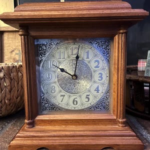 Puede incluir: Un reloj de chimenea de madera con una esfera cuadrada y una esfera decorativa plateada y blanca. El reloj tiene una base rectangular y una parte superior decorativa. Las manecillas del reloj indican la hora. El reloj es de madera.
