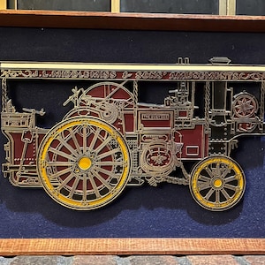 Puede incluir: Obra de arte enmarcada que presenta una ilustración detallada de una máquina de vapor vintage. La obra es principalmente burdeos y dorada, con el texto "J.L. Middlemiss & Sons' Amusements" en la parte superior. Se encuentra sobre un fondo azul oscuro dentro de un marco de madera.