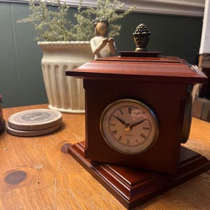 May include: A small, rectangular, wooden clock with a round clock face. The clock has a dark brown finish and a decorative finial on top. The clock face has Roman numerals and gold-colored hands. The clock sits on a wooden base.