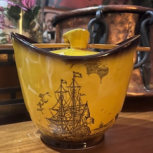 May include: A yellow ceramic sugar bowl with a dark brown rim and base, featuring a black line drawing of a sailing ship on the front. The bowl includes a matching yellow lid with a central handle and a thin wooden spoon resting across the top.