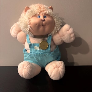 May include: A plush doll with light skin, blue eyes, and a brown nose, featuring a head of shaggy cream-colored yarn hair. The doll wears light blue denim-style overalls with a small white label on the chest and a brown collar with a gold medallion.