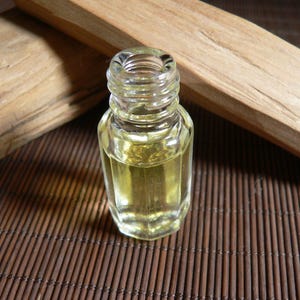 May include: A small, clear glass bottle containing a yellow liquid, possibly essential oil, is positioned on a brown bamboo mat. Two pieces of light-coloured wood are in the background. The bottle has a hexagonal shape and a threaded neck.