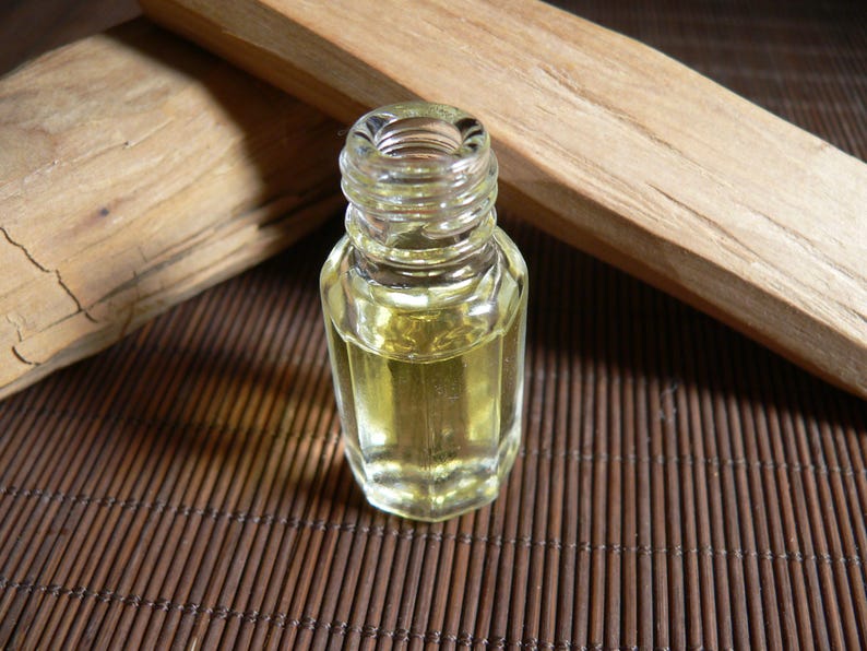 May include: A small, clear glass bottle containing a yellow liquid, possibly essential oil, is positioned on a brown bamboo mat. Two pieces of light-coloured wood are in the background. The bottle has a hexagonal shape and a threaded neck.