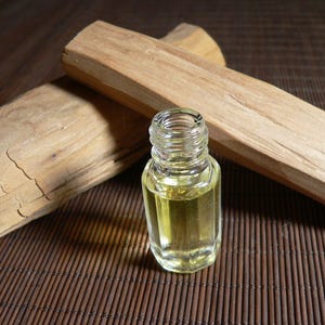 May include: A small, clear glass bottle containing yellow liquid, next to two light brown sandalwood logs. The logs have a textured surface and visible grain. The items are on a brown bamboo mat.