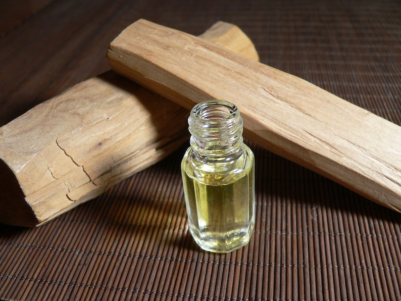 May include: A small, clear glass bottle containing yellow liquid, next to two light brown sandalwood logs. The logs have a textured surface and visible grain. The items are on a brown bamboo mat.