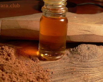 Op de afbeelding: Een klein glazen flesje gevuld met een goudbruine vloeistof, dat op een houten oppervlak ligt met bruin poeder en een houten stok. De fles heeft een doorzichtige glazen stop.