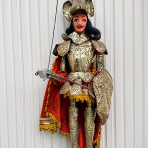 Könnte beinhalten: Eine Marionette als Ritter in einer goldfarbenen Metallrüstung mit detaillierten Gravuren. Die Figur trägt einen leuchtend roten Umhang mit Goldfransen, einen Helm mit Federbusch und hält ein Metallschwert sowie einen Schild. Schwarzes Haar.