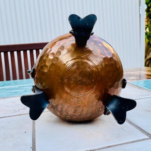 Puede incluir: Un cuenco decorativo de cobre martillado con detalles en metal negro. El cuenco tiene forma redonda con una superficie texturizada y tres formas de aleta. La palabra "gekro" es visible en la parte inferior del cuenco.