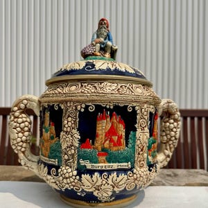 May include: A detailed ceramic stein with a relief design. The stein features a castle scene with red roofs, green trees, and the text "Burg Eltz. Mosel." A small figurine sits atop the lid. The handles are shaped like grape clusters.