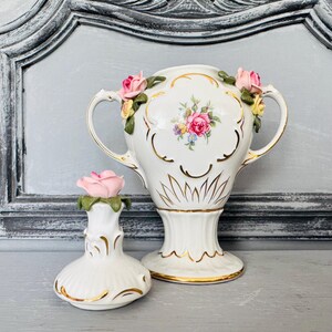Może przedstawiać: Biały porcelanowy wazon z dwoma uchwytami oraz pasujący do niego mały flakonik na perfumy, oba ozdobione złotymi wykończeniami oraz trójwymiarowymi różowymi i żółtymi różami. Wazon posiada centralny bukiet kwiatów i żłobkowaną podstawę.