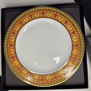 Puede incluir: Un plato de cerámica blanca con un borde decorativo. El borde presenta un patrón repetido de diseños florales dorados, rojos y amarillos. El plato está colocado en una caja negra.