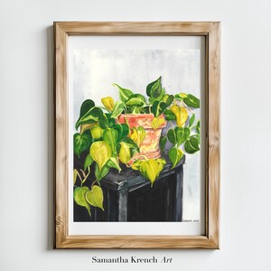 May include: Watercolor painting of a potted plant with green and yellow leaves. The plant is in a terracotta pot and is sitting on a black table. The painting is framed in a light wood frame.