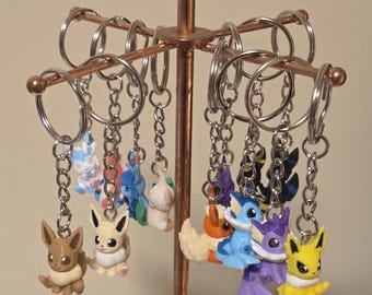 May include: A collection of small, colourful plastic character keychains hanging from a copper-coloured metal display stand. Each keychain features a different stylised creature with distinct colours, including brown, blue, yellow, and purple, attached to a silver metal ring and chain.