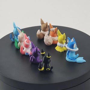 May include: A collection of colorful, small figurines of various creatures, including blue, pink, purple, yellow, brown, and black characters. The figurines are arranged on a black circular surface.