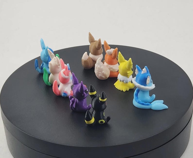 May include: A collection of colorful, small figurines of various creatures, including blue, pink, purple, yellow, brown, and black characters. The figurines are arranged on a black circular surface.