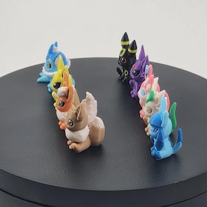 May include: A collection of colorful figurines, each depicting a unique creature. The figurines are arranged in two rows on a black circular surface. The creatures are various colors, including blue, yellow, brown, purple, pink, and green.
