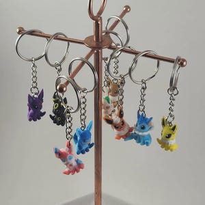 May include: A collection of colorful character keychains hanging from a copper-colored display stand. The keychains feature various cartoon figures in shades of purple, black, blue, red, white, orange, and yellow. Each keychain has a silver-colored chain and ring.