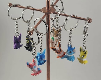 May include: A collection of colorful character keychains hanging from a copper-colored display stand. The keychains feature various cartoon figures in shades of purple, black, blue, red, white, orange, and yellow. Each keychain has a silver-colored chain and ring.