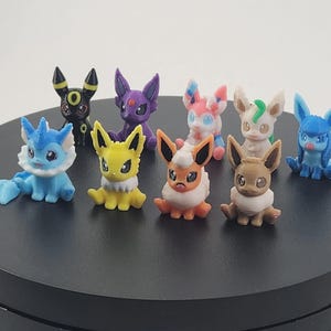 May include: A collection of nine colorful, small figurines of various Pok&eacute;mon characters. The figurines are in different colors, including blue, yellow, purple, and brown. They are arranged on a black circular surface.