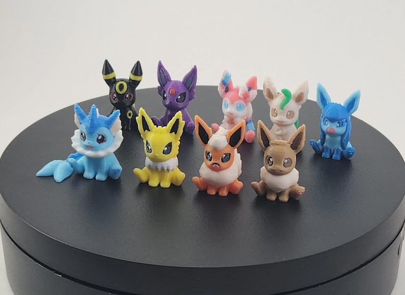 May include: A collection of nine colorful, small figurines of various Pok&eacute;mon characters. The figurines are in different colors, including blue, yellow, purple, and brown. They are arranged on a black circular surface.