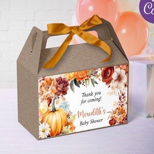 Può includere: Una scatola regalo marrone con un nastro dorato e un design floreale con zucche e foglie autunnali. La scatola ha un'etichetta che dice "Grazie per essere venuto! Baby Shower di Meredith".