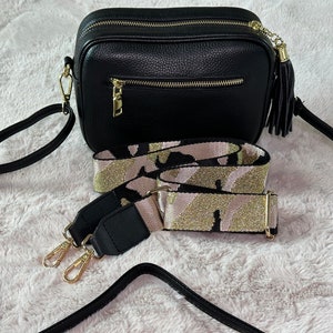 May include: A black leather crossbody bag with a gold zipper and tassel. The bag comes with a black and gold patterned adjustable strap.