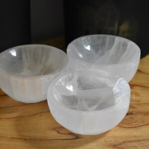 Peut inclure: Trois petits bols en sélénite blancs et translucides sont disposés sur une surface en bois. Les bols ont une forme lisse et arrondie. Deux objets noirs sont en arrière-plan. Idéal pour la décoration.