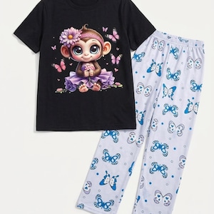 May include: A black short-sleeved t-shirt featuring a graphic of a cartoon monkey wearing a purple skirt and a flower headband, surrounded by pink butterflies. The set includes white pajama pants with a repeating pattern of blue butterflies.