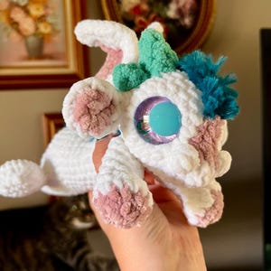 May include: A handmade white crochet creature with pink and teal accents. The plush toy has large, iridescent blue eyes and tufts of teal and blue yarn. The creature's paws and ears are detailed with pink yarn.