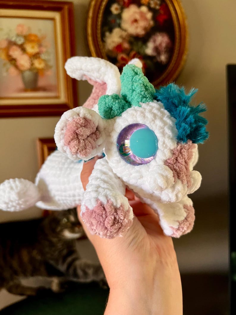 May include: A handmade white crochet creature with pink and teal accents. The plush toy has large, iridescent blue eyes and tufts of teal and blue yarn. The creature's paws and ears are detailed with pink yarn.