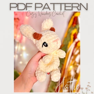 Könnte beinhalten: Ein gehäkeltes Amigurumi-Spielzeug in Form eines Hasen mit braunen Ohren und einem weißen und cremefarbenen Körper. Der Hase wird von einer Hand gehalten und der Hintergrund ist verschwommen. Der Text "Cozy Wonders Crochet" und "Hattie" ist auf dem Bild zu sehen.