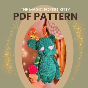 May include: A green crocheted stuffed animal with orange and gold antlers. The animal has a sad expression and is holding a pink and orange pom-pom. The text "THE MAGIC FOREST KITTY PDF PATTERN" is at the top of the image.