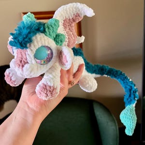 May include: A handmade crochet dragon plushie in white, teal, pink, and blue. The dragon has large, glittery eyes, a fluffy mane, and a long tail. The plushie is soft and cuddly, perfect for a child's toy or a unique gift.