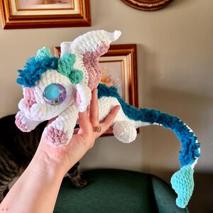 May include: A handmade crochet dragon in white, pink, and blue. The dragon has large, blue eyes and a long, curved tail. The dragon is being held up by a hand.