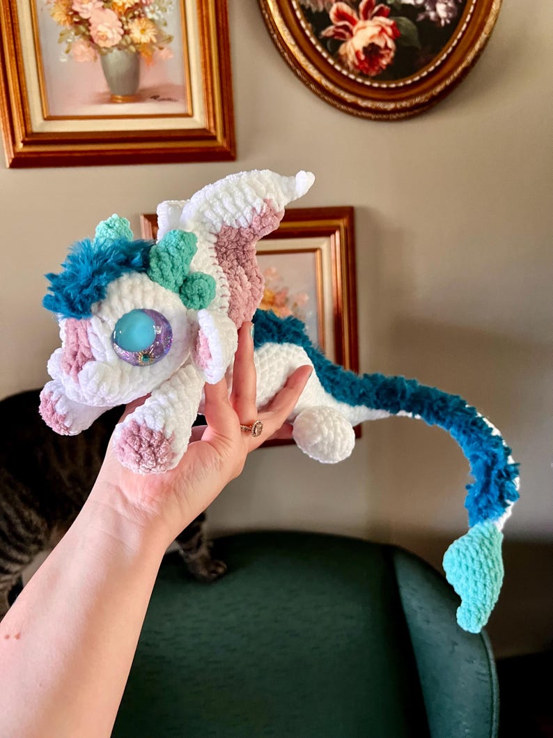 May include: A handmade crochet dragon in white, pink, and blue. The dragon has large, blue eyes and a long, curved tail. The dragon is being held up by a hand.